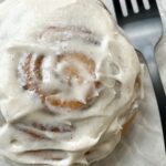 A soft and fluffy cinnamon roll smothered in spiced maple cream cheese frosting and served on a glass plate.