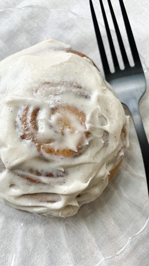 A soft and fluffy cinnamon roll smothered in spiced maple cream cheese frosting and served on a glass plate.